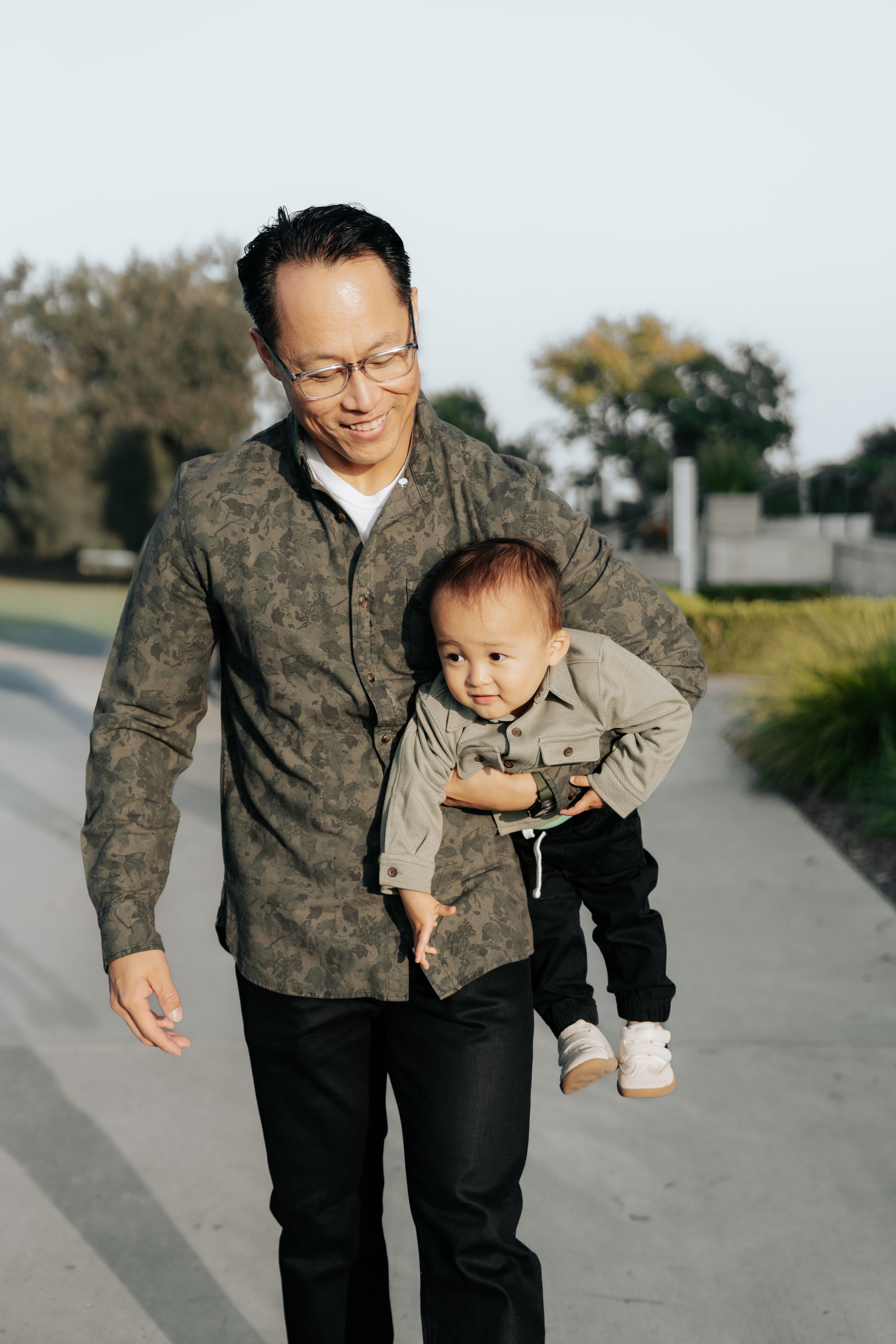 A man smiling while carrying a young child on his hip, both dressed in casual clothing, walking along a path with greenery in the background.